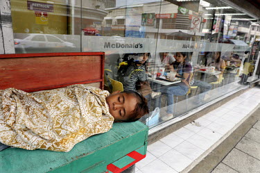 A street vendor's child sleeps on a bench beside a McDonald's fast food outlet as the diners look on.
