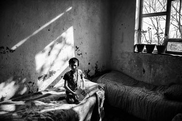 A boy sits on a bed in the light of a large window at the Kabul Orphanage.