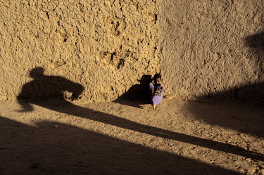 A small child sits against a wall, with a shadow of a person elongated by the sun in the town of Gao, located on the Niger River.