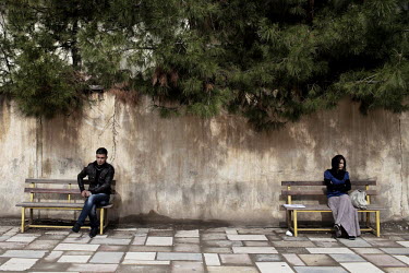 A young man sits on one bench, while a young woman sits on another one, in the yard of the Duhok Polytechnic University.