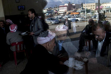 Elderly men play cards at a cafe with the street reflected in the window.