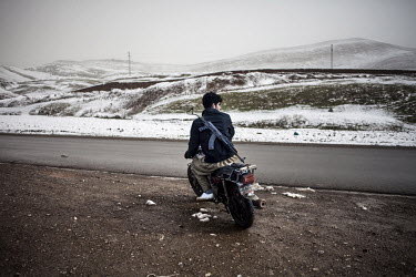 An armed security guard patrols near the Iranian border on a motorcycle, with snow covered hills in the background.