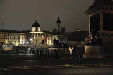 A woman takes a picture of a child sitting on one of the lion sculptures on Trafalgar Square next to Nelson's column with the National Gallery visible in the background.