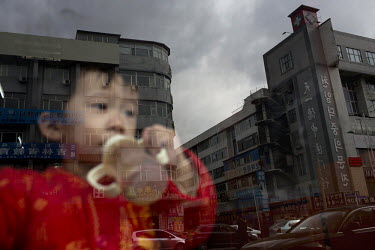 A child, reflected in a window pane, drinks from a bottle as he looks out on to a street. The buildings outside sport signage in both Korean & Chinese scripts.