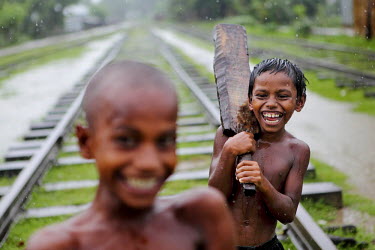 A boy carrying a hand carved cricket bat stands behind another boy on a railway track in the rain.