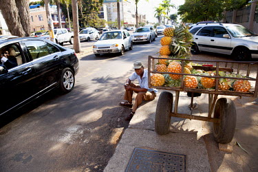 A man driving a Mercedes-Benz car passes a pinapple vendor sitting at the roadside.