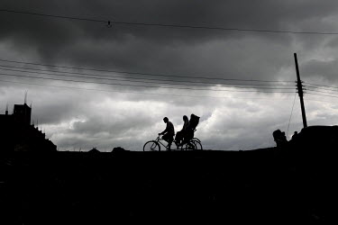 A rickshaw driver and his passenger are silhouetted against a stormy sky.