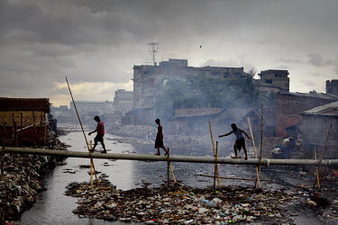 Three men cross a bamboo bridge over a river that is clogged with rubbish.