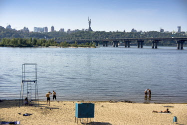 People on a sandy beach beside the Dnipro River. In the background is the Moscow Bridge and on the horizon is the Mother of the Fatherland, a monument comemorating those honoured with the title 'Hero...