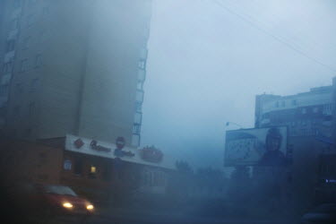 A car drives past a Soviet era tower block on a misty evening with a propaganda poster showing a child visible through the mist.