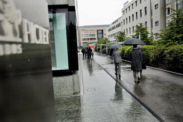 Businessmen, shelter from the rain beneath umbrellas, as they head to work in a newly developed banking quarter in the Acasias district. Pictet & Cie, private bankers are to the left and UBS at the en...