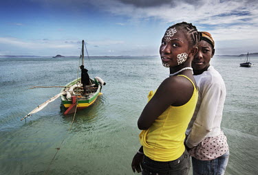 Two women, one who has decorated her face with a geometric design, watches a sail boat race from the shore.
