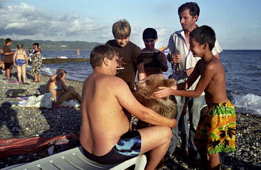 A tourist holds onto a bear which is being led by a hawker who offers tourists the opportunity to have their picture taken with the bear. Children pet the bear enthusiastically.