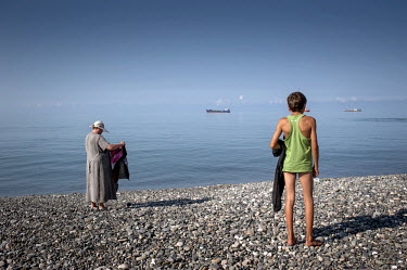 A woman and a boy stand on the pebble beach at Sukhumi. A cargo ship is visible in the distance.