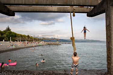 A girls jumps into the water from the remnants of a concrete structure on Sukhumi beach. A boy swings gets ready to swing into the water on a rope suspended from the concrete structure and bathers enj...