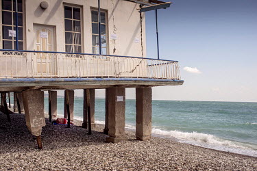 A woman lies in the shade below a beach hut built on stilts at Sukhumi beach. The doors and walls of the building have 'Danger' signs on them.