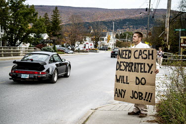 A Porche car passes a man standing at the roadside holding a sign that reads: 'NEED CASH DESPERATELY, WILL DO ANY JOB!!!" Manchester Village is a wealthy town in New England.
