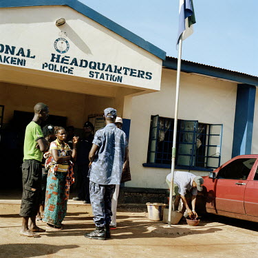 People petition a police officer in the entrance to the central police station in Makeni, whilst a man prepares to clean the officer in command's red Mercedez Benz car.