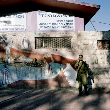 An Israeli Defence Force (IDF) soldier stands near a large mural painted by settlers. It depicts their version of Hebron's history from ancient times to: 'Liberation, Return, and Rebuilding'...'1967:...
