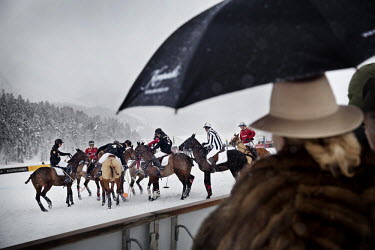 Dressed in warm furs and sheltering beneath an umbrella, a spectator watches a mele of horses and riders during the final match at the 30th Polo on Snow World Cup. The game between Team Cartier and Te...