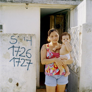 A woman stands outside her home, baby in arms. The wall next to her is marked for demolition with the initials of the municipal housing secretary and an identifying number. At 115 years old, Providenc...