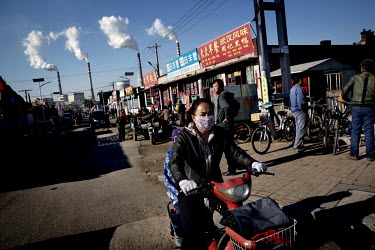 A woman, wearing a surgical mask, rides her scooter past a row of shops. In the background smoke stacks at the Datong No. 2 coal-fired power plant rise on the horizon.