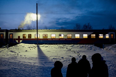 A view of the Matvei Mudrov train at dusk with a group of people standing in the snow outside.   The Matvei Mudrov train is a medical train operated by Russian Railways along the course of the Baikal...