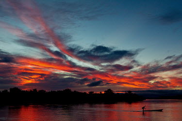 A man punts a dugout canoe across the Congo River. Above him a dramatic sunset has coloured the sky and water bright orange.