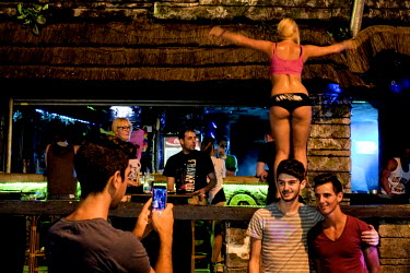 Two young men have their picture taken in front of a bar, making sure the partially clad young female dancer standing behind them is included in the snap.