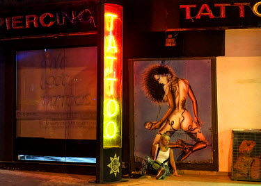 A drunk tourist slumped against a poster of a naked, tattooed woman at a tattoo parlour.
