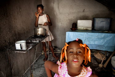 Judeline, a 'restavek' cooking food while the six-year-old daughter of her host, Bubu, plays. 'Restaveks' are children whose impoverished parents have indentured them into domestic labour in the hope...