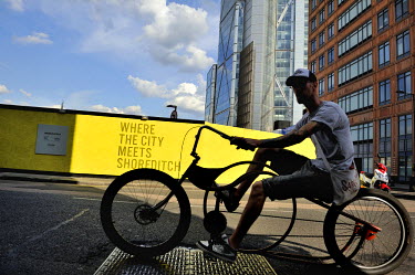 Low slung cyclist cyclist passing 'Where the City meets Shoreditch', a slogan on hoarding at a development site between Shoredicth and the City in the East End.