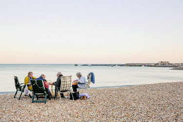 A group of elderly men and women enjoy a picnic on the shingle beach, beside the Cobb or the harbour at Lyme Regis.