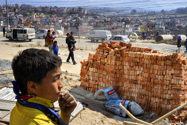On the side of the Ring Road, which not long ago marked the edge of Kathmandu, Nepalese people walk through clouds of dust brought on by construction activity.  In the background a dense cluster of ne...
