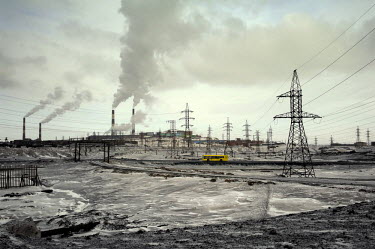 A yellow bus drives through a grey, brown and black landscape with industrial infrastructure and smokestacks in the Greater Norilsk area.  Greater Norilsk consists of three settlements: Norilsk, Talna...