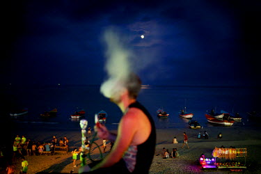 From a terrace above the beach a young man smokes a shisha (sheesha, narghile, hookah) while watching the moon and the party coming to life. The full moon party on Haad Rin Beach on the island of Ko P...