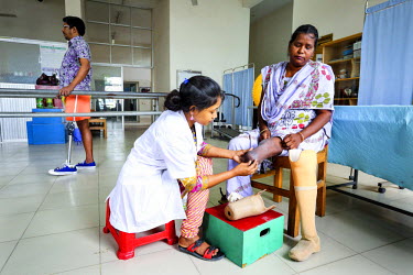 Ripa Akter (P & O assistant) helps Anowara Begum (40) fix her artificial limb below her knee. She lost her leg in a road accident in 2011. In the background Imrahim Morshed is practicing walking with...
