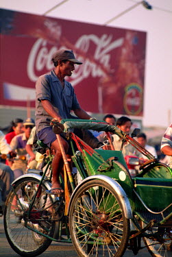A bicycle rickshaw rider waits at a road junction in front of a Coca-Cola billboard advertisement.