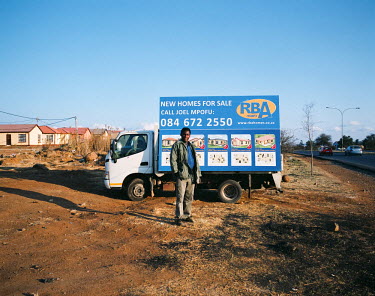A man stands in front of his small van which bears an advertising board offering ^new homes^ in Devland Park, a new housing development to the southeast of Soweto. Joel comes to this spot every day wi...