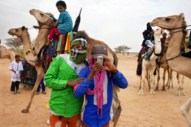 A boy takes a picture with a mobile phone while behind camels and riders look on.