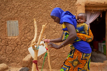 Madina (30), with her baby daughter Aissatou (one) strapped to her back, prepares to wash her hands using a water container on a frame designed for maximum hygiene.