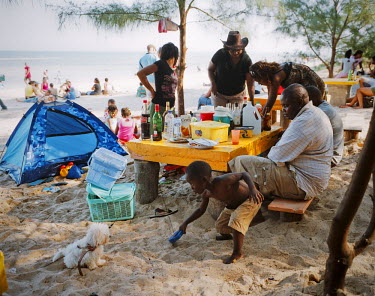 A family has a picnic on a beach near Maputo. A boy plays with the family dog while adults relax in the shade.