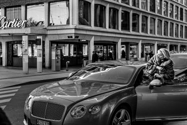 A woman, dressed in fur and clutching a Chanel purse, enters a Bentley car parked near a Cartier shop on Rue du Rhone, a thoroughfare reknown for its luxury brand shops.