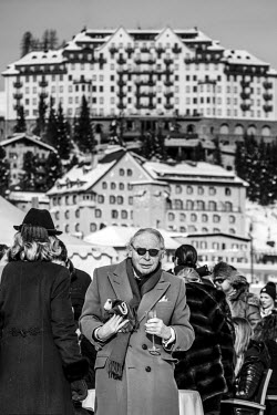 An elderly man holding a glass of champagne in his hand during a pause in the action at the annual Snowpolo tournament. In the background is the Carlton Hotel.