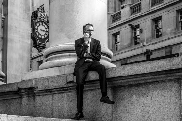 A businessman uses a mobile phone while sitting on a wall outside the Royal Exchange building in the City of London.