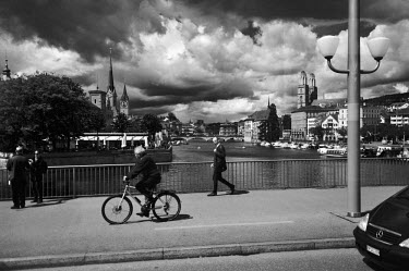 A cyclist and pedestrians crossing over the Limmat River with the city behind. On the right bank are the twin domes of the Grossmunster and on the left bank is the Fraumunster.