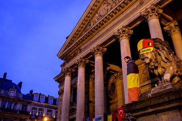A man wrapped in a Belgian flag stands beside a similarly attired statue of a lion at the impromptu shrine to victims of the 22 March terrorist attacks that has grown up in front of the Bourse (the St...