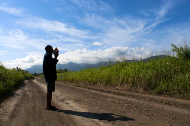 Antipas Kiriwang, standing on a rural road, blows a conch shell (a traditional form of communication and warning in Papuan traditional societies).