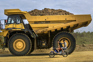 Two miners riding a motorbikeare dwarfed by an enormous dump truck carrying waste from a commercial jade mine.