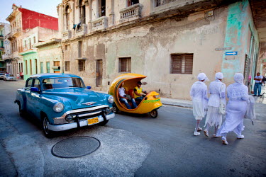 A group of Santeria initiates cross a road in front of a classic American car and a three wheeled taxi. After their initiation santeros must wear white for a year, they may not drink alcohol and must...
